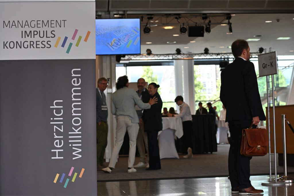 Conference lobby with attendees networking; a tall banner on the left reads 'Management Impuls Kongress' and a man in a suit stands on the right.
