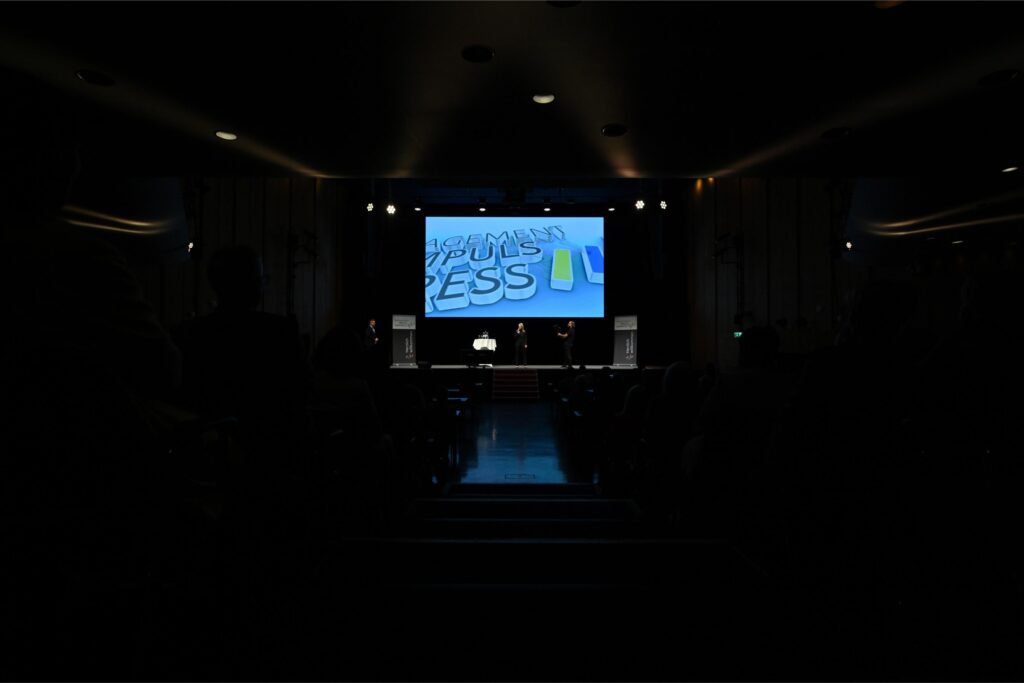Dimly lit auditorium with a bright blue presentation on a large screen; several people stand on the stage in front of the screen, audience silhouettes in the foreground.