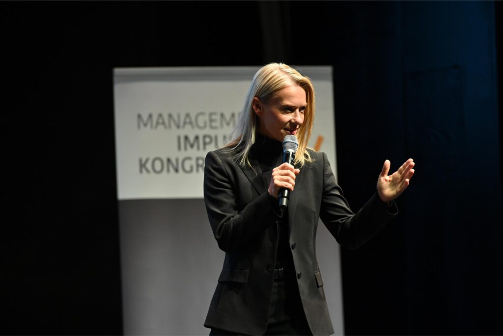 Female speaker on stage holding a microphone and gesturing with one hand, dark background and a partial sign behind her.