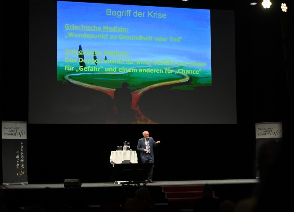 Presenter standing at a podium on a dark stage, a large slide in blue behind him with German text about crisis and medicine hints (theme slide).