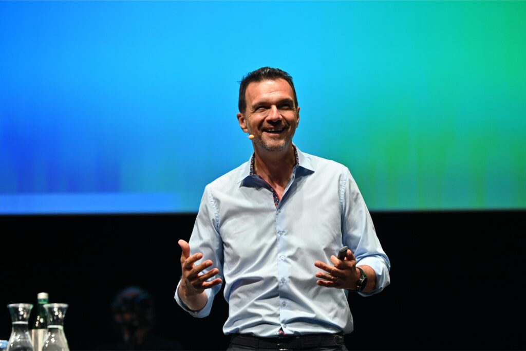 Smiling man on stage giving a talk, wearing a light blue shirt and headset mic, gesturing with hands against a blue‑green backdrop.