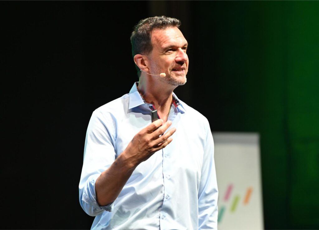Speaker on stage wearing a light blue shirt and headset microphone, gesturing with his hands during a talk.