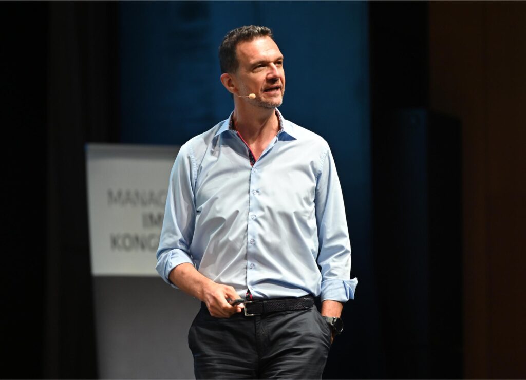 Male speaker on stage wearing a light blue shirt with a headset microphone, gesturing while giving a talk.
