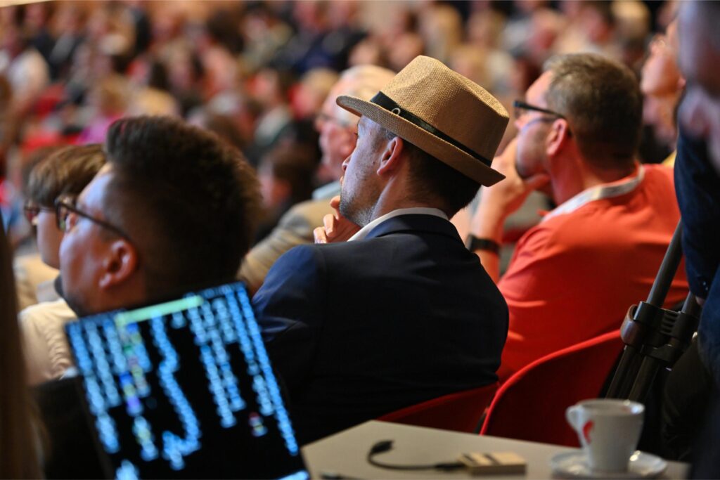 Audience at a conference or talk; man in a tan fedora in focus, others blurred in background.