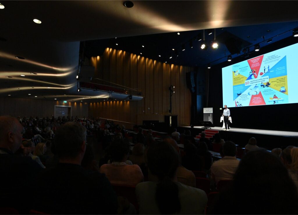 Speaker presenting on a lit stage to a darkened auditorium, with a large colorful infographic slide on the screen