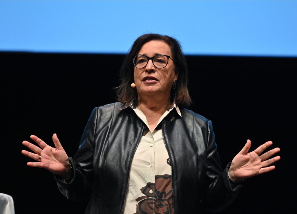 Woman in a black leather jacket and glasses speaks on stage, hands raised in gesture against a dark backdrop.