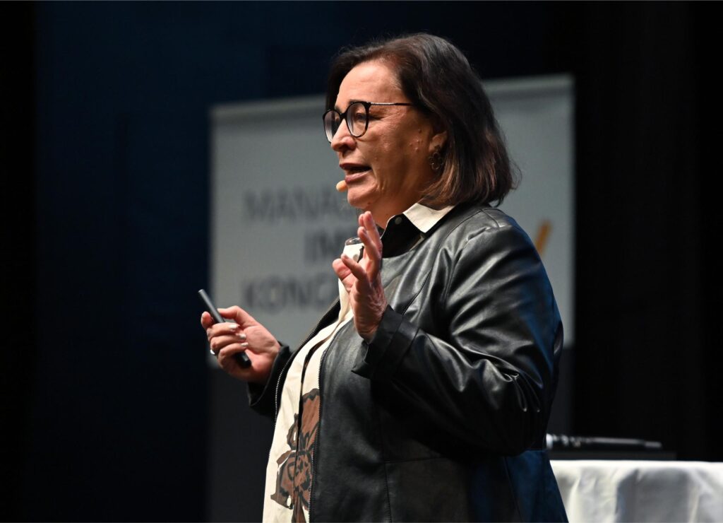 Speaker on stage wearing glasses and a black leather jacket, gesturing with her hands while talking into a headset microphone