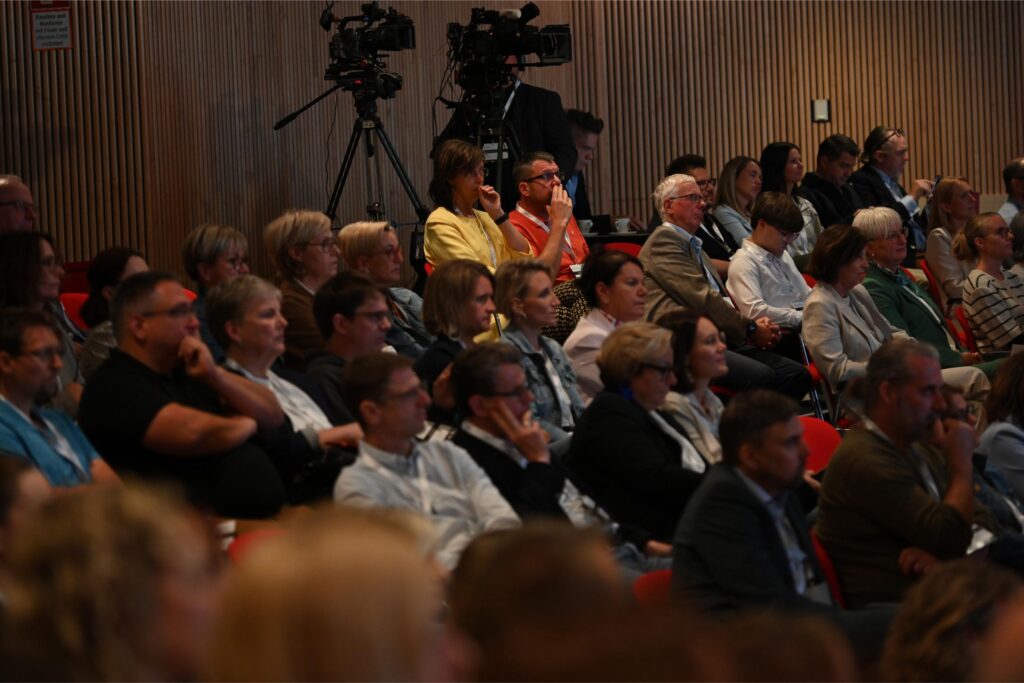 Audience seated in a wood-paneled conference hall, with two camera operators on tripods at the back of the room.