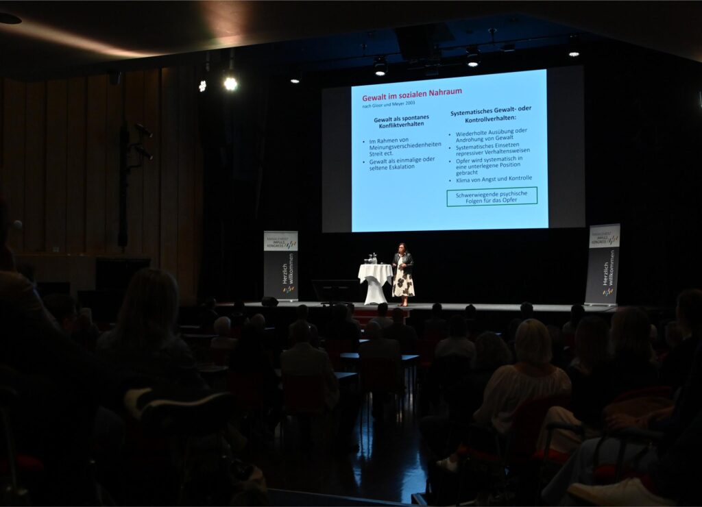 Speaker on stage presenting slides about violence in the social environment to an audience in a dark auditorium.