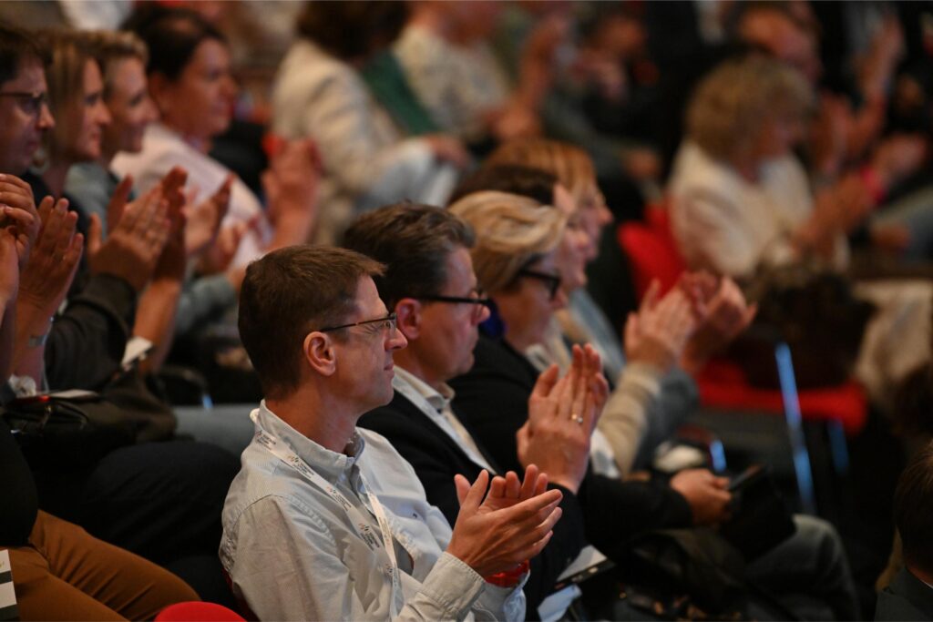 Audience members in a conference or lecture hall clapping and watching a speaker.