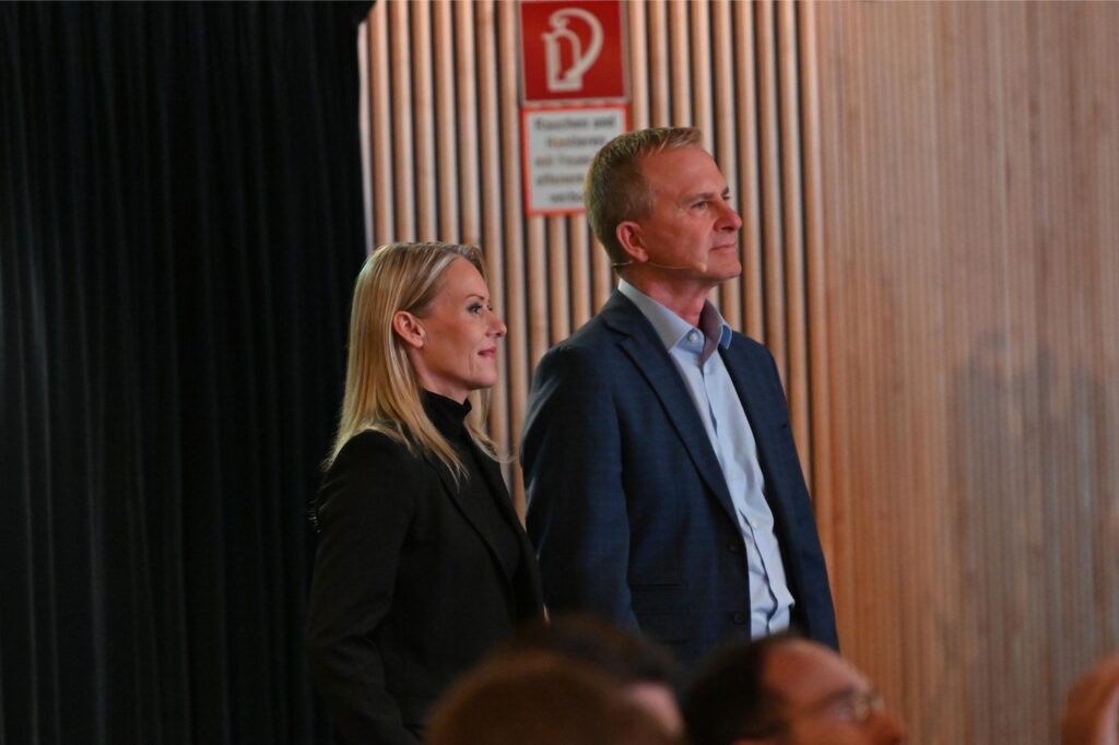 Two adults in business attire stand on a stage, side by side, looking toward the audience amidst wood-paneled backdrops and a red safety sign in the background.