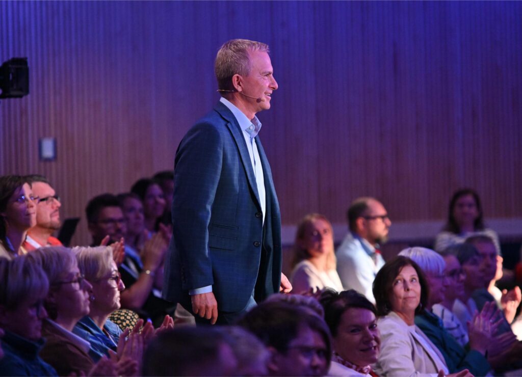Man in a blue blazer stands on stage with a headset mic, addressing an audience at an event.