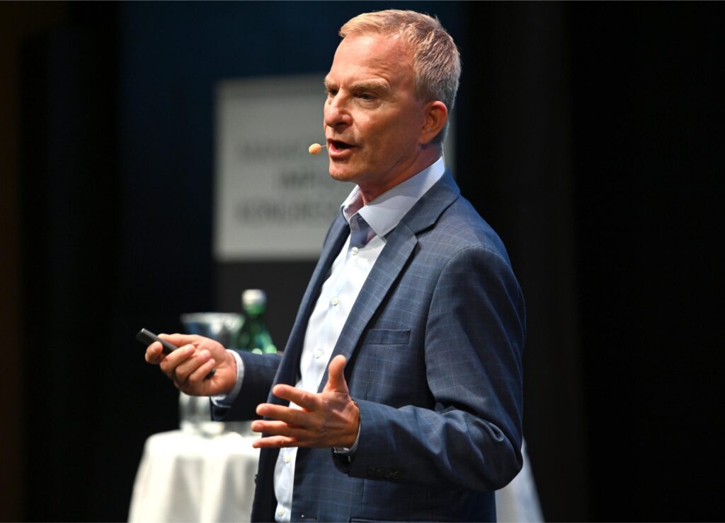 Male presenter in a blue checkered suit speaks on stage with a headset microphone, gesturing with his hands.