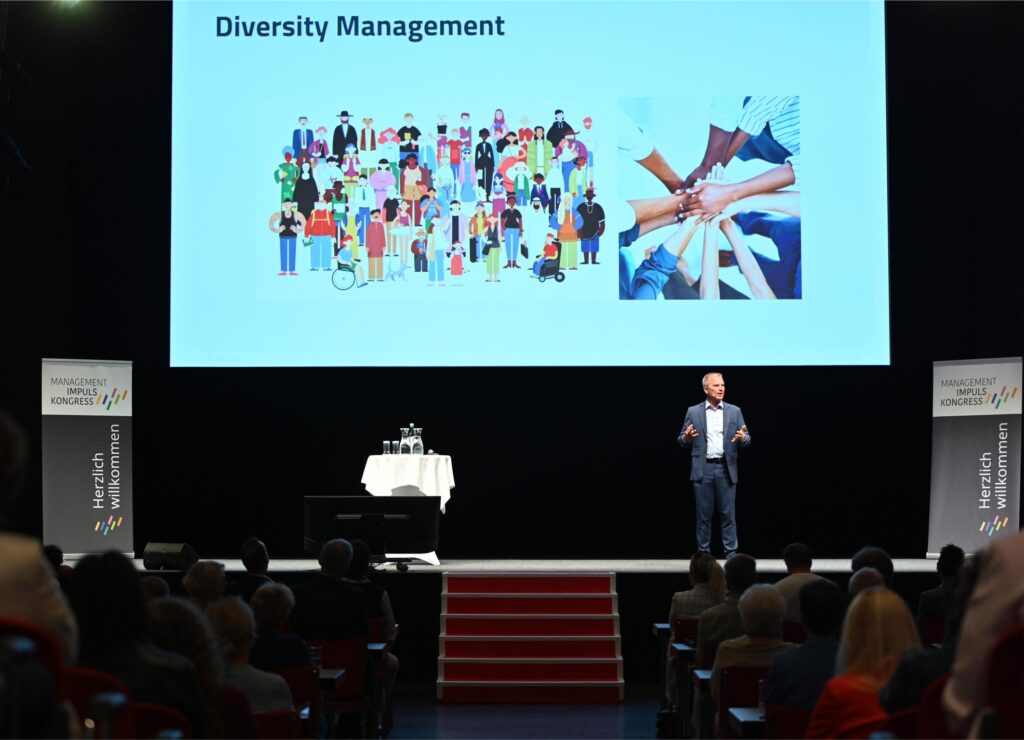 Speaker presenting on a stage with a large screen reading 'Diversity Management' and images of diverse people and hands in a group hug to symbolize teamwork, in a conference setting.