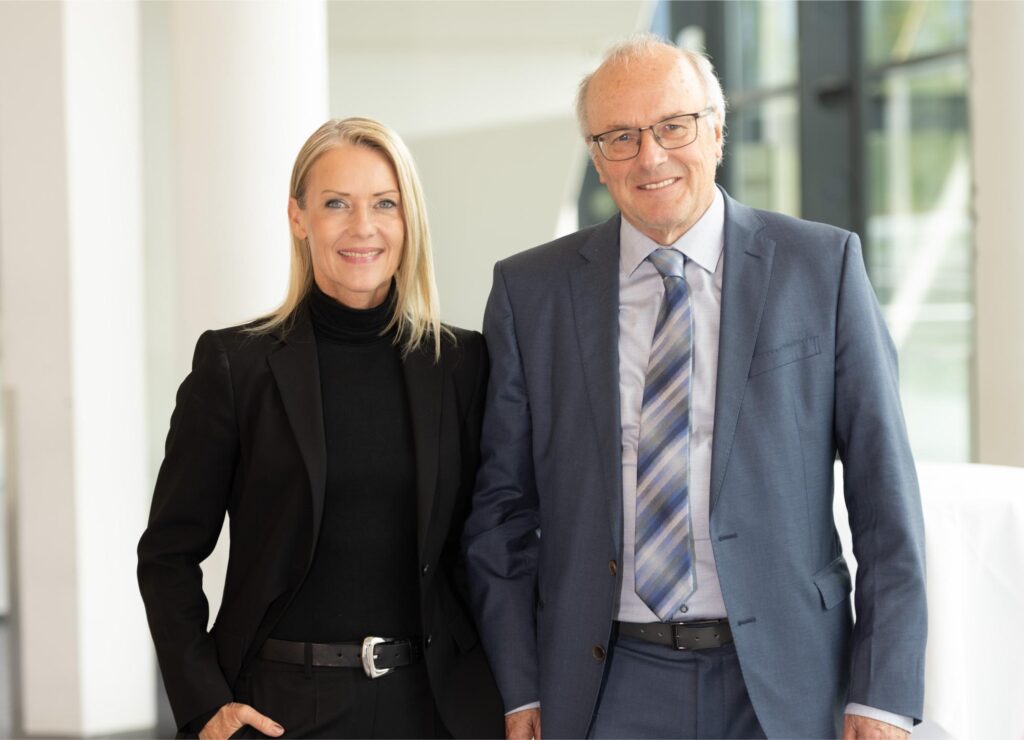 Two professionals in business attire pose together in a bright, modern hallway, smiling at the camera.