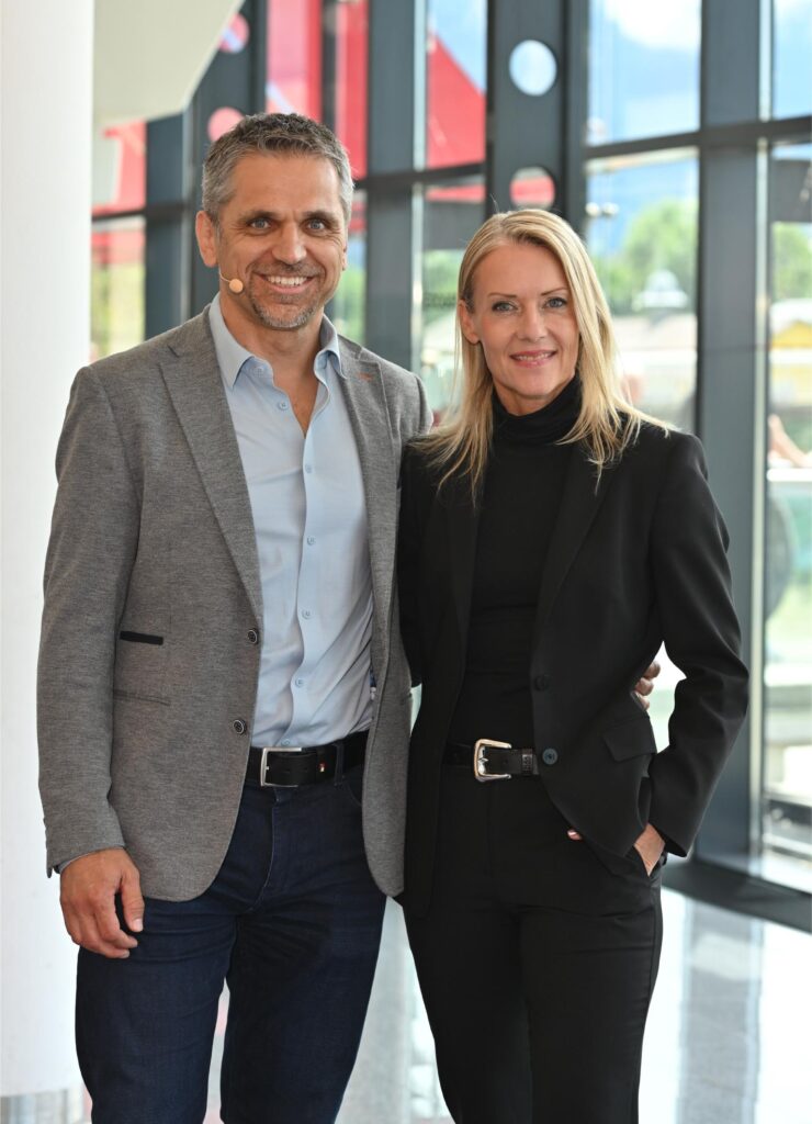 Two smiling professionals, a man in a gray blazer and a woman in a black suit, posing together in a bright modern lobby.