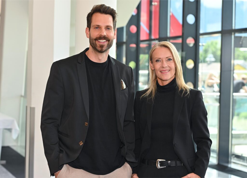 Two professionally dressed coworkers pose and smile in a bright, modern lobby area.
