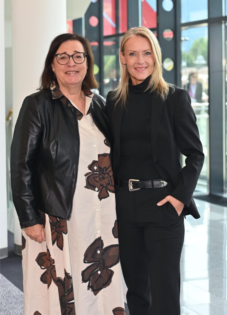 Two women stand together indoors, smiling at the camera; left in a beige floral dress with a black leather jacket, right in a black blazer and pants with a belt.