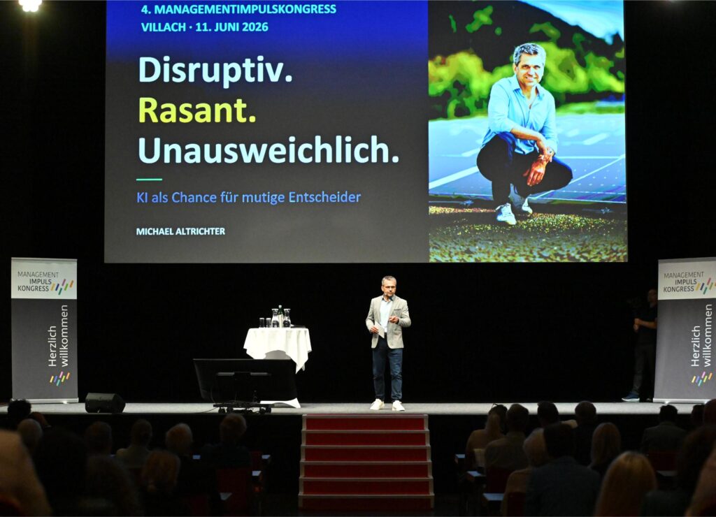 Speaker on stage at a management congress with a large screen showing the German headline 'Disruptiv. Rasant. Unausweichlich.' and 'Managementimpuls Kongress' in the background.