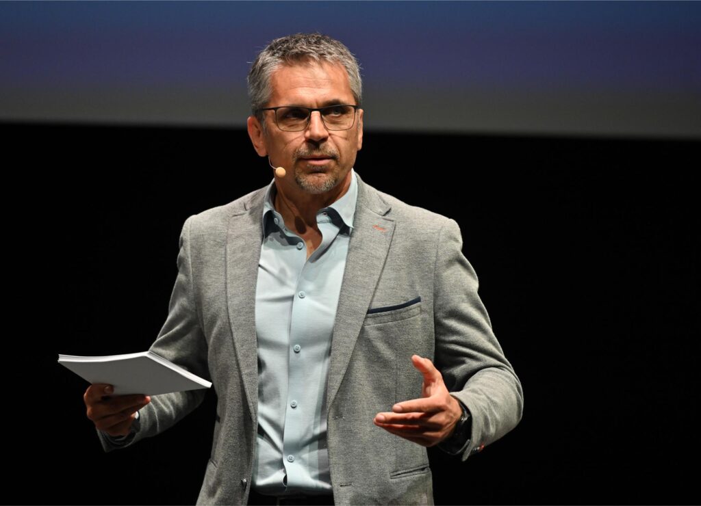 Man in a gray blazer and light blue shirt delivering a presentation on stage, holding papers.