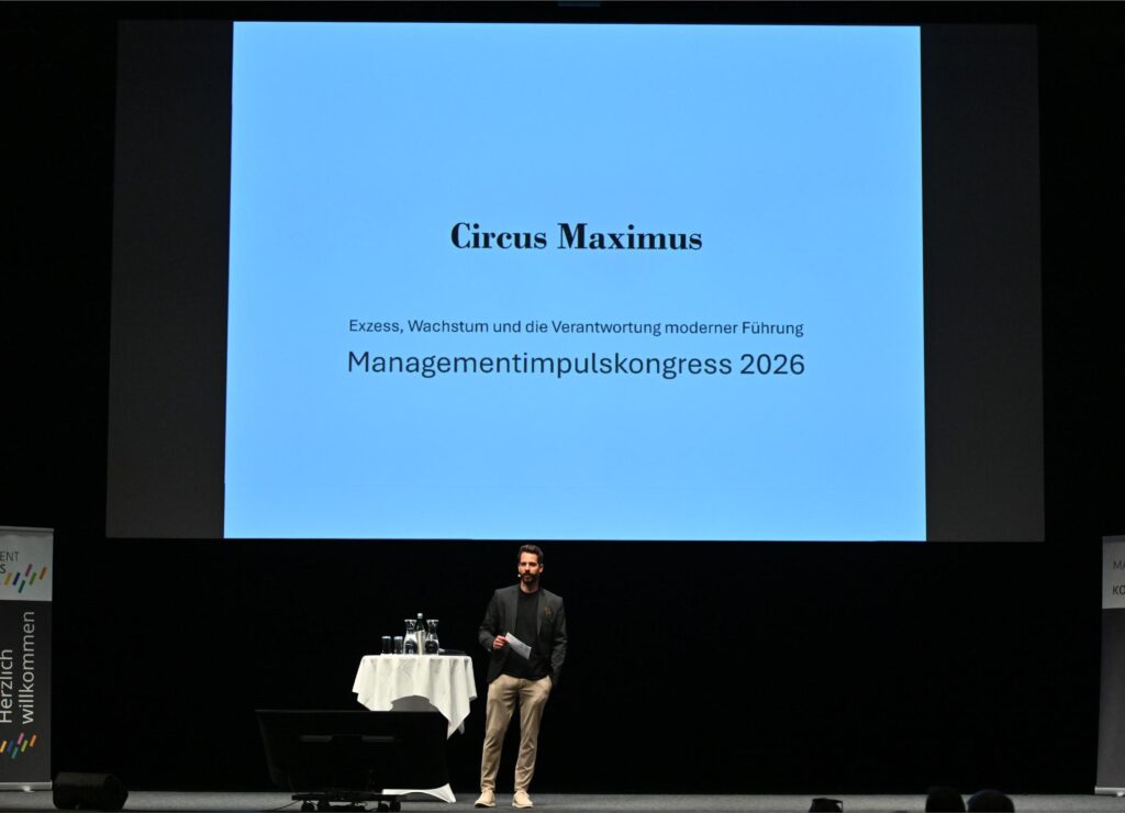 Male presenter on stage in front of a blue slide reading 'Circus Maximus' and 'Managementimpulskongress 2026'.