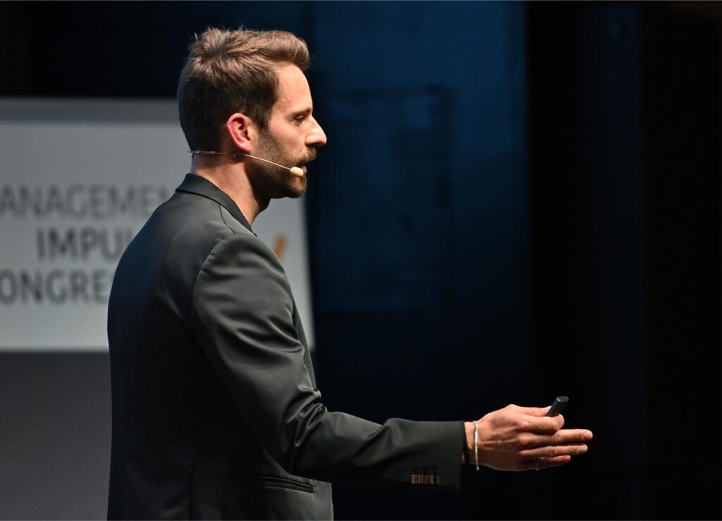Side-profile of a man in a dark suit giving a presentation on a stage, wearing a headset microphone