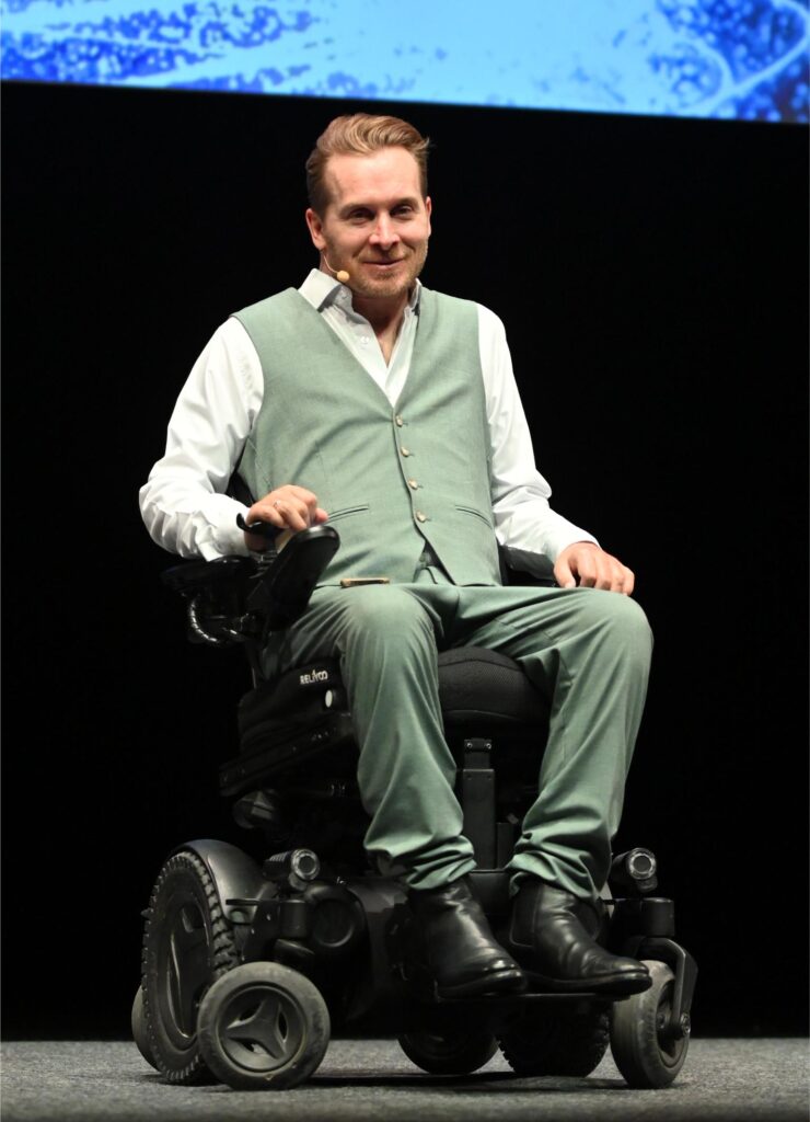 Male presenter in a mint-green suit sitting in a motorized wheelchair on stage, wearing a headset microphone.