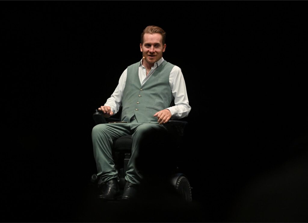 Speaker in a green vest and white shirt sits in a wheelchair on a dark stage, wearing a headset microphone and gesturing with his hands as he speaks into the audience.