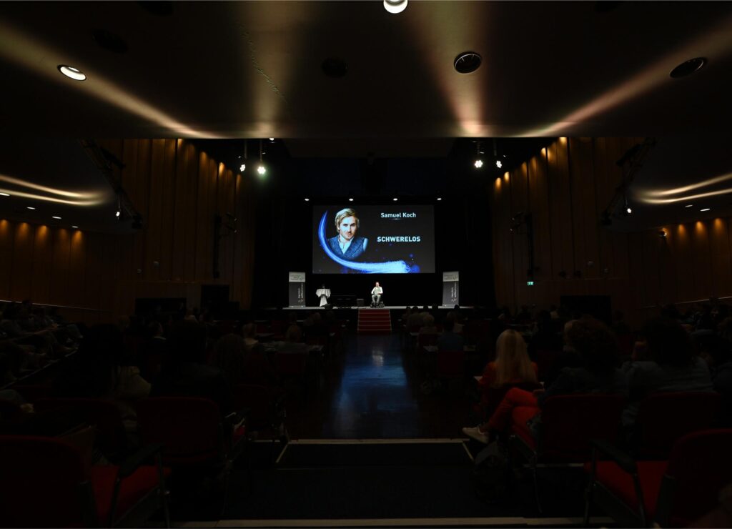 Dim auditorium with a stage and a large screen showing a presenter; screen reads 'Samuel Koch' and 'Schwerelos.'