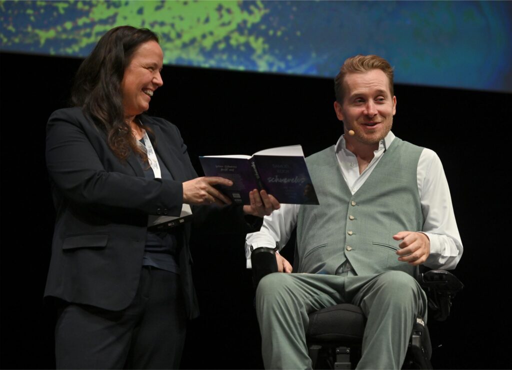 Woman in a dark blazer hands a booklet to a smiling man in a light gray vest seated in a wheelchair on stage.