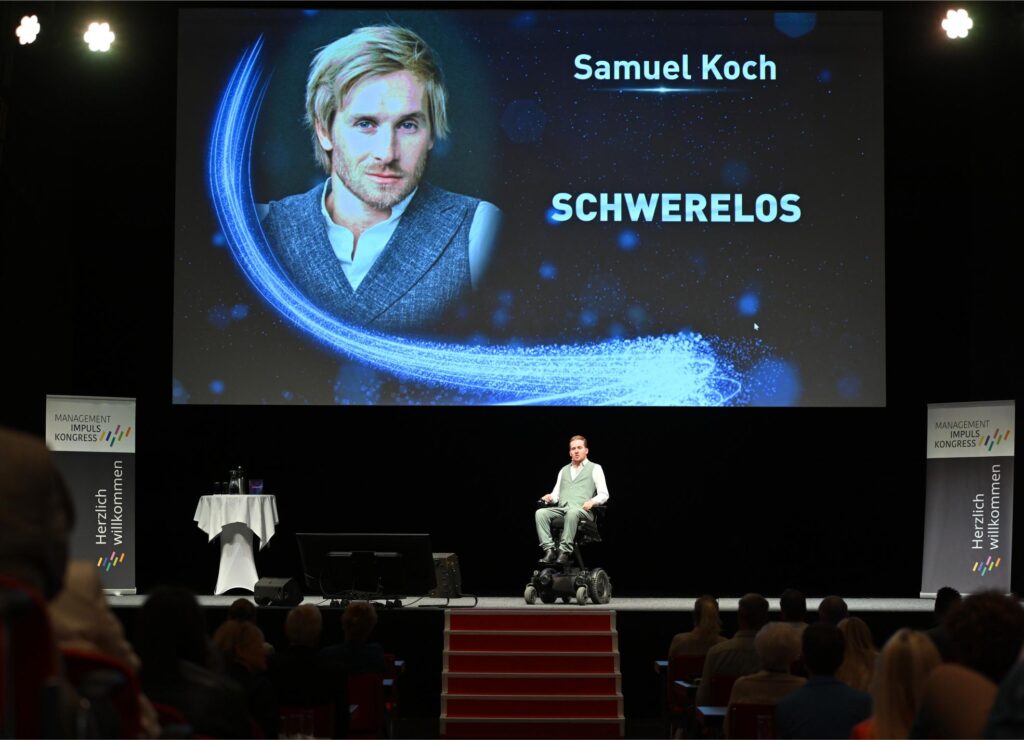 Stage presentation: man in a wheelchair on a lit stage, with a large screen showing 'Samuel Koch' and 'SCHWERelos' and blue cosmic graphics in the background.