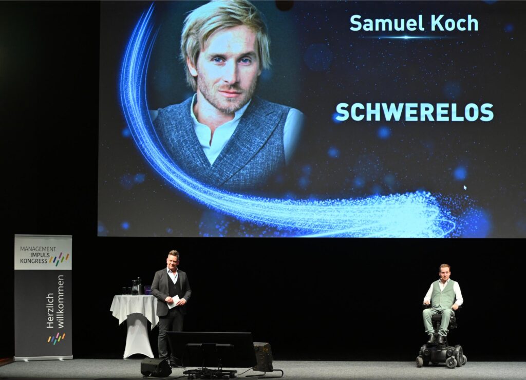Stage presentation: man in suit at podium on a dark stage, large screen behind shows portrait and the words 'Samuel Koch' and 'SCHWERelos' with blue light effects, and a man in a wheelchair on the right.