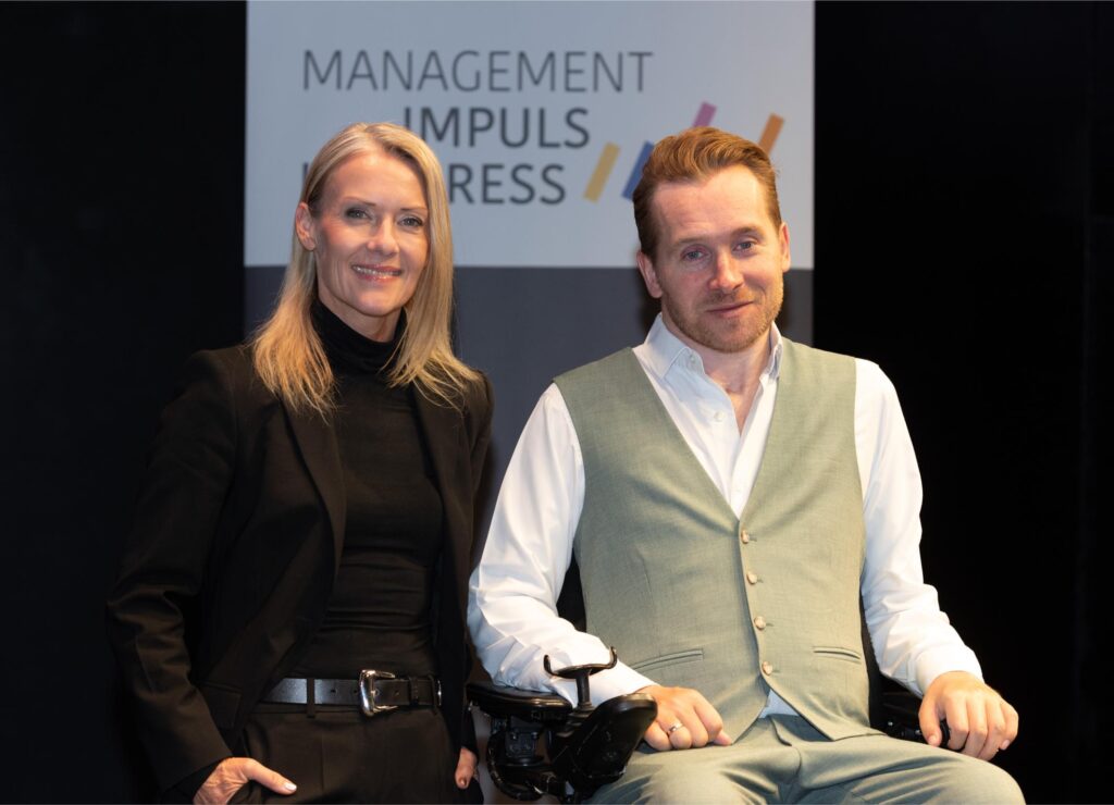 Two professionals posing together at an event, a woman in black outfit on the left and a man in a light gray vest on the right, smiling in front of a sign.